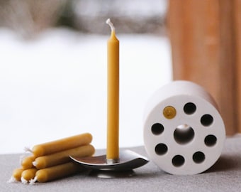 May include: A collection of beeswax candles and candle holders. A tall, yellow candle stands in a silver holder, with a stack of smaller candles nearby. A white, cylindrical candle holder with multiple holes is also present.