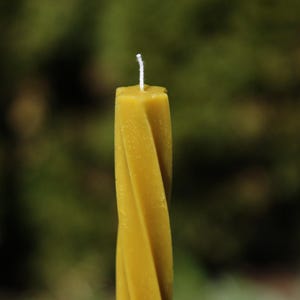 以下が含まれることがあります： ねじれたデザインの黄色の蜜蝋キャンドルのクローズアップ。キャンドルは白い芯とテクスチャ加工された表面を持っています。背景はぼやけた緑色です。