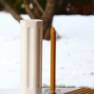 Puede incluir: Un molde de vela hexagonal blanco con una sola vela de cera de abeja amarilla y un grupo de cinco velas de cera de abeja amarilla.