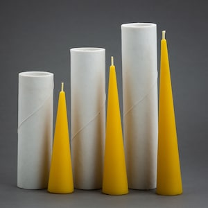 Puede incluir: Cuatro tubos de cartón blanco con cuatro velas cónicas amarillas. Las velas son de diferentes tamaños y están dispuestas en fila de la más pequeña a la más grande.