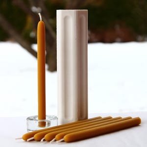 Pode incluir: Um conjunto de seis velas de cera de abelha, incluindo uma vela alta única em um porta-velas de vidro e cinco velas mais curtas. As velas são de cor amarelo dourado e estão dispostas sobre uma superfície branca. Também está incluído um molde retangular branco para fazer velas.