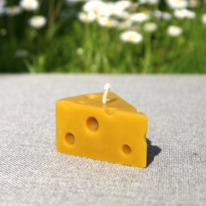 Puede incluir: Una vela amarilla con forma de queso con tres agujeros y una mecha blanca. La vela está sobre una superficie gris con un fondo borroso de hierba verde y flores blancas. Un artículo decorativo único.