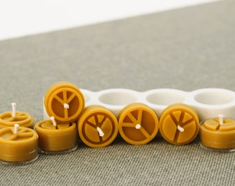 May include: A set of eight small, round, yellow beeswax candles with a peace sign design. The candles are arranged in a row on a brown fabric surface.