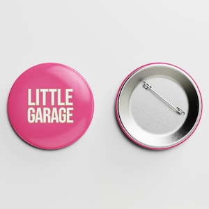 May include: A pink button with the text "LITTLE GARAGE" in white lettering. The back of the button is silver with a safety pin.