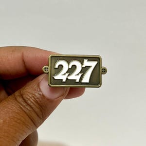 May include: A gold-colored metal pin with the number 227 in white raised lettering. The pin has a rectangular shape with rounded corners.