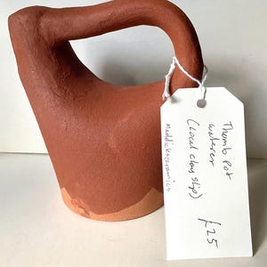 May include: A terracotta-coloured ceramic waterer with a curved handle and a wide base. A white rectangular paper tag is attached with string, featuring handwritten text that reads: Thumb Pot waterer, Local clay slip, maddicksceramics, £25.