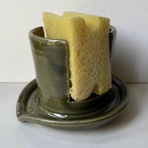 Peut inclure: Un porte-éponge en céramique fait à la main, dans des tons de vert, avec un égouttoir intégré. Il contient deux éponges jaunes. Le design est simple et fonctionnel, idéal pour la cuisine ou la salle de bain.