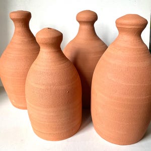 Op de afbeelding: Vier terracotta-kleurige vazen met een afgeronde body en een smalle hals. De vazen hebben een getextureerd oppervlak en een eenvoudig, klassiek ontwerp. De vazen zijn dicht bij elkaar geplaatst en tonen hun vergelijkbare vormen en maten.