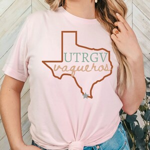 Pu&ograve; includere: T-shirt rosa chiaro con un contorno marrone dello stato del Texas. La maglietta mostra il testo "UTRGV vaqueros" in verde e beige. Una stella grigia si trova nella parte inferiore del contorno del Texas. Girocollo.