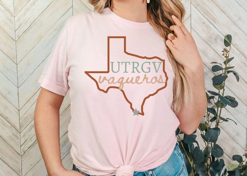 Pu&ograve; includere: T-shirt rosa chiaro con un contorno marrone dello stato del Texas. La maglietta mostra il testo "UTRGV vaqueros" in verde e beige. Una stella grigia si trova nella parte inferiore del contorno del Texas. Girocollo.