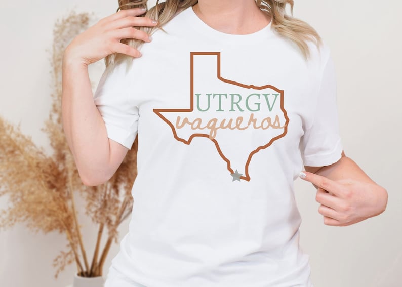 Pu&ograve; includere: T-shirt bianco con un contorno marrone dello stato del Texas e la scritta "UTRGV vaqueros" in verde e beige. Una stella grigia si trova nella parte inferiore del contorno del Texas. La maglietta &egrave; girocollo.