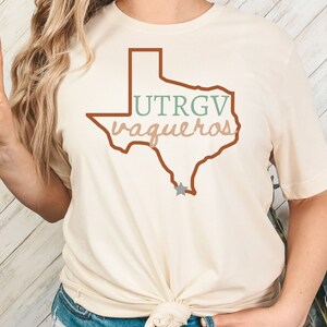 Pu&ograve; includere: T-shirt color crema con un contorno marrone dello stato del Texas. La maglietta mostra la scritta "UTRGV Vaqueros" in verde e beige, con una stella grigia. Una maglietta casual per tutti i giorni.
