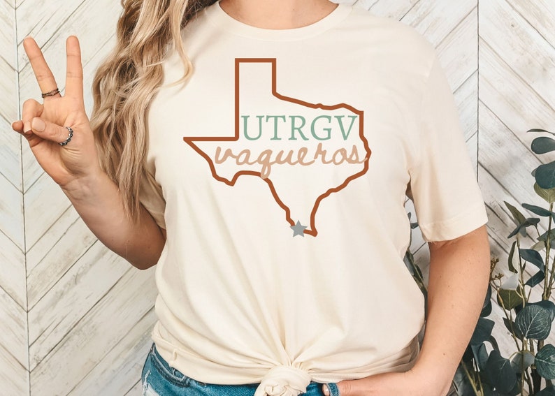 Pu&ograve; includere: T-shirt color crema con un contorno marrone dello stato del Texas. La maglietta mostra la scritta "UTRGV Vaqueros" in verde e beige, con una stella grigia. Una maglietta casual per tutti i giorni.