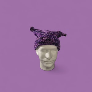 May include: A purple and black crocheted hat with pointed tips, displayed on a white mannequin head against a solid purple background. The hat's texture is visible, showcasing the yarn's colour variations.