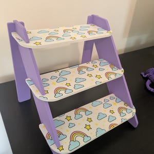 May include: A three-tiered, lilac-coloured wooden shelf unit. The shelves feature a pattern of rainbows, clouds, and stars on a white background. The unit is designed in a ladder style.