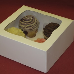 May include: White cardboard cupcake box with clear plastic windows showing four cupcakes. The cupcakes are decorated with purple, white, yellow, and pink frosting.
