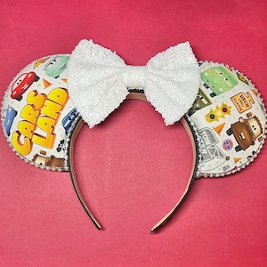 May include: A Cars Land themed headband with a white sequined bow. The ears feature a Cars Land print with images of cars and the Cars Land logo. The headband is trimmed with silver beads and has a brown band.