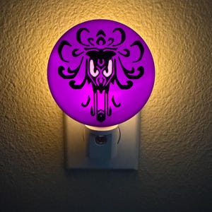 May include: A circular night light featuring a vibrant purple glow with a prominent black damask-style wallpaper pattern printed on the surface. The light is plugged into a standard wall outlet, emitting a soft illumination from behind the disc.