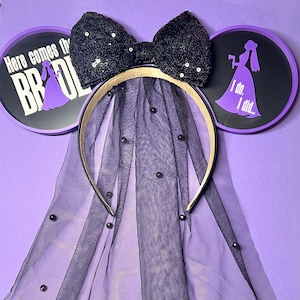 May include: A black sequined bow headband with a sheer black veil adorned with small black beads. The headband features two circular purple and black ear accents with text and a bride silhouette. Text on the ears reads "Here comes the BRIDE" and "I do. I did..."