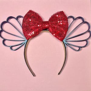 May include: A headband featuring a large, red sequined bow and blue seashell-shaped accents. The headband has a neutral-toned band and is set against a pink background. The design is reminiscent of a mermaid or aquatic theme.