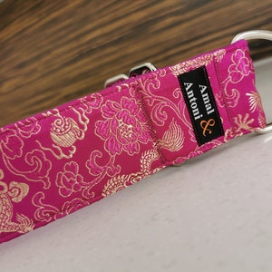 May include: A pink and gold patterned dog collar with a silver buckle and D-ring. The collar has a black label with the text "Amal & Antoni" in white.
