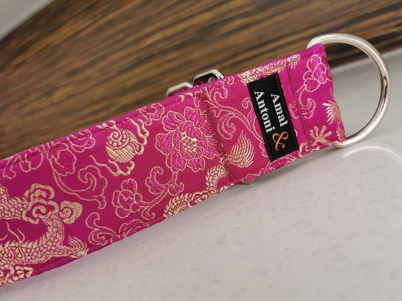 May include: A pink and gold patterned dog collar with a silver buckle and D-ring. The collar has a black label with the text "Amal & Antoni" in white.