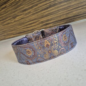 May include: A dog collar with a paisley pattern in shades of blue, gold, and burgundy. The collar has a soft, blue interior and silver-colored metal hardware. The collar is designed for a dog.
