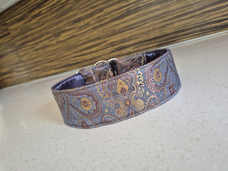 May include: A dog collar with a paisley pattern in shades of blue, gold, and burgundy. The collar has a soft, blue interior and silver-colored metal hardware. The collar is designed for a dog.