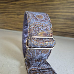 May include: A dog collar with a blue and gold paisley pattern. The collar has a silver metal buckle and D-ring. The fabric has a detailed floral design.