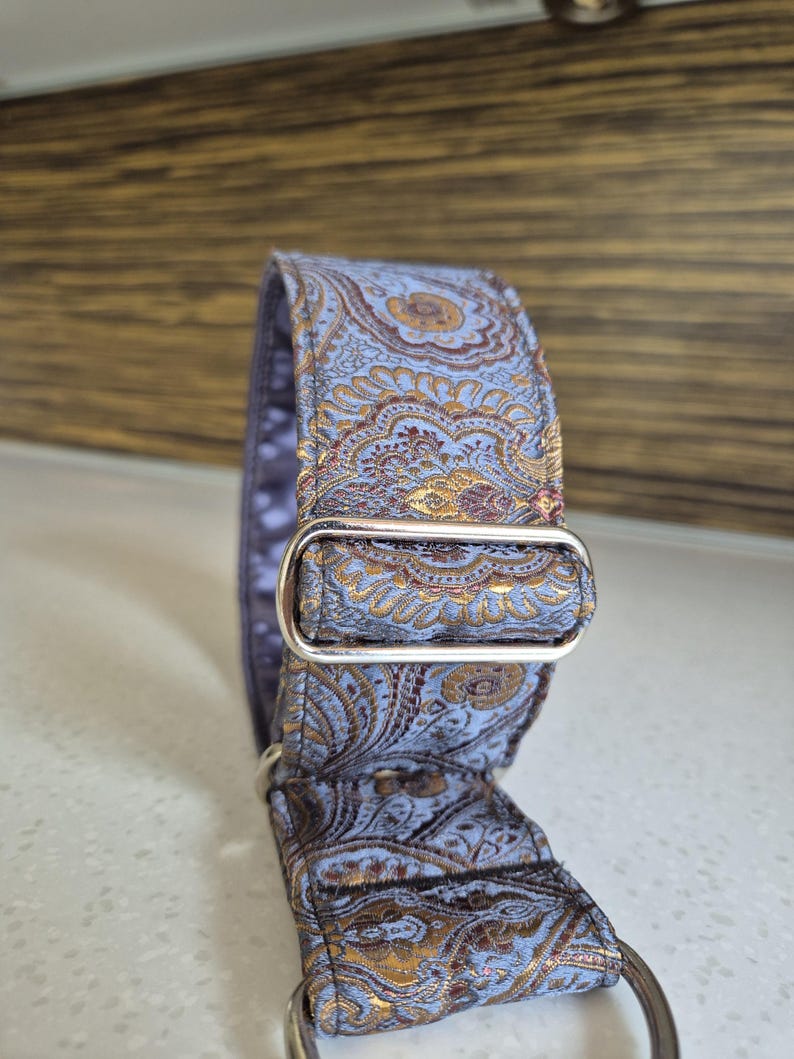 May include: A dog collar with a blue and gold paisley pattern. The collar has a silver metal buckle and D-ring. The fabric has a detailed floral design.