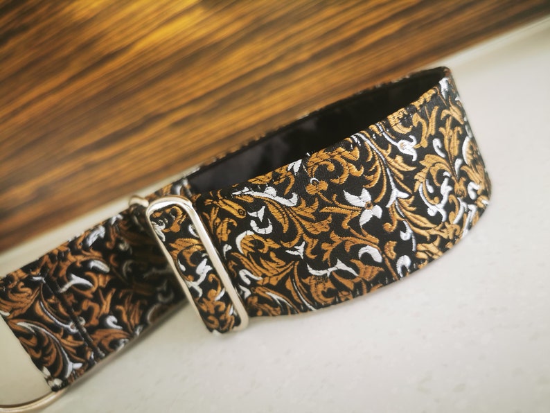 Puede incluir: Un collar de perro con estampado negro, dorado y blanco con una hebilla plateada.