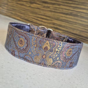 May include: A wide dog collar with a paisley pattern in shades of blue, gold, and burgundy. The collar has a soft, padded interior and silver-colored metal hardware. The fabric has a subtle sheen.