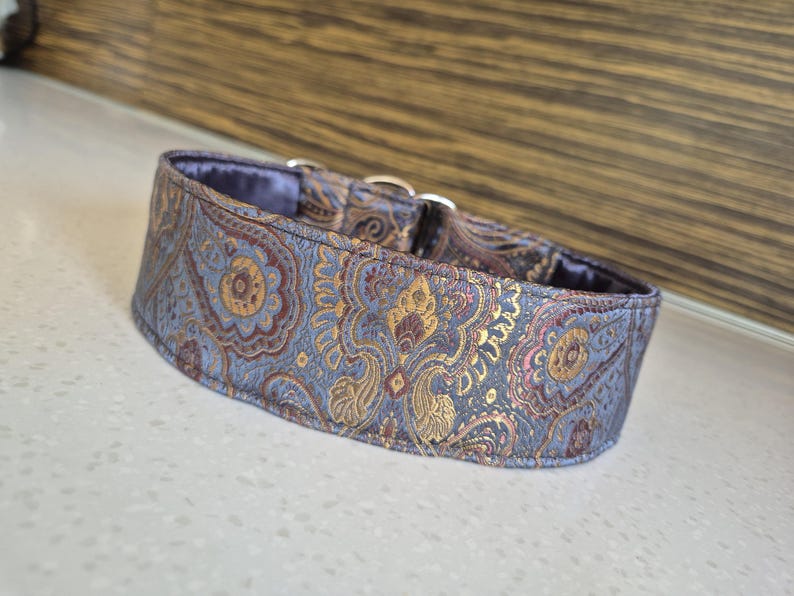 May include: A wide dog collar with a paisley pattern in shades of blue, gold, and burgundy. The collar has a soft, padded interior and silver-colored metal hardware. The fabric has a subtle sheen.