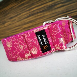 May include: A pink dog collar with a gold floral pattern and a silver buckle. The collar has a black label with the text "Amal & Antoni" in white.