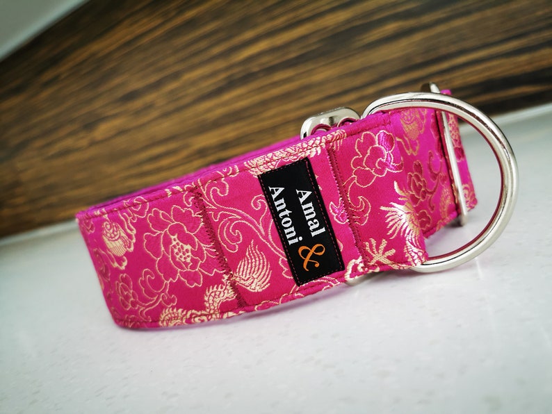 May include: A pink dog collar with a gold floral pattern and a silver buckle. The collar has a black label with the text "Amal & Antoni" in white.