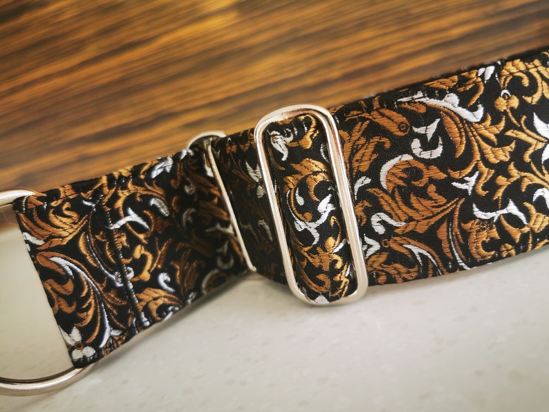 Puede incluir: Un collar de perro con estampado negro, dorado y blanco con una hebilla de metal plateado.