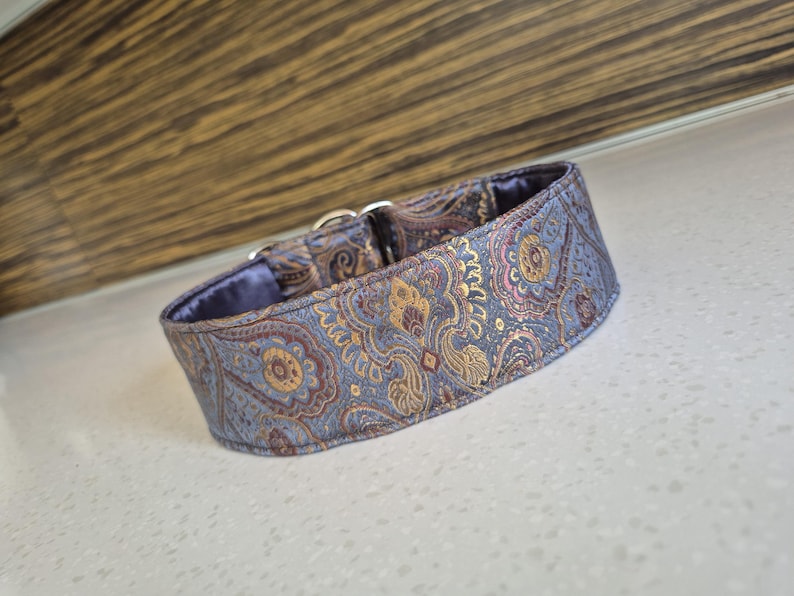 May include: A decorative dog collar with a paisley pattern in shades of blue, gold, and burgundy. The collar has a soft, purple lining and a silver metal buckle. This collar is designed for pets.