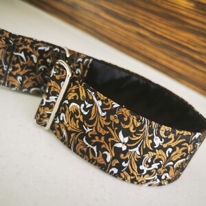 Puede incluir: Un collar de perro con estampado negro, dorado y blanco con un anillo en D plateado.