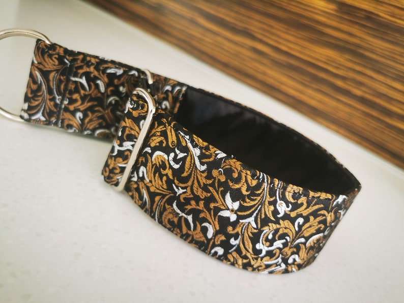 Puede incluir: Un collar de perro con estampado negro, dorado y blanco con un anillo en D plateado.