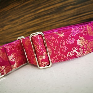 May include: A pink and gold dog collar with a dragon pattern. The collar has a silver buckle and a D-ring.