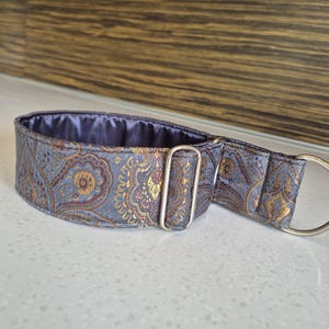 May include: A grey and gold paisley dog collar with a silver metal D-ring and buckle. The collar has a padded interior and is designed for comfort and style. The pattern includes gold and burgundy accents.