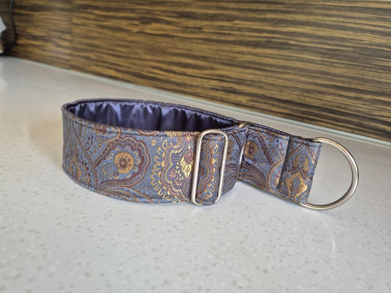 May include: A grey and gold paisley dog collar with a silver metal D-ring and buckle. The collar has a padded interior and is designed for comfort and style. The pattern includes gold and burgundy accents.
