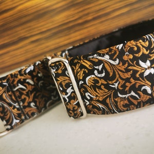 Puede incluir: Un collar para perro negro y dorado con un dise&ntilde;o floral y una hebilla plateada. El collar est&aacute; hecho de tela.