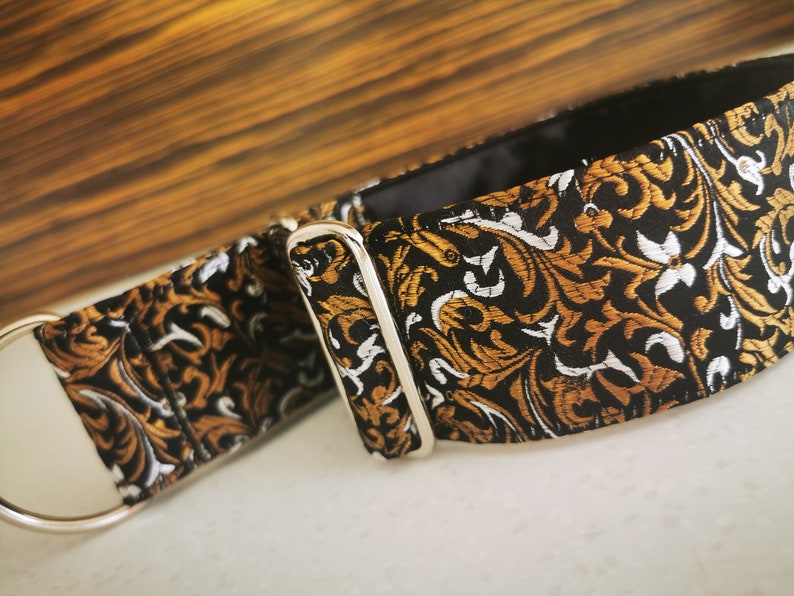 Puede incluir: Un collar para perro negro y dorado con un dise&ntilde;o floral y una hebilla plateada. El collar est&aacute; hecho de tela.