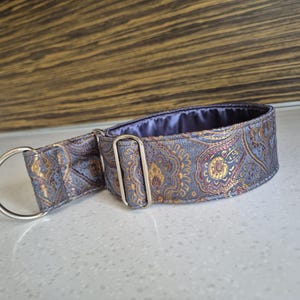 May include: A patterned dog collar with a silver metal D-ring and buckle. The collar features a paisley design in shades of blue, gold, and purple. The interior lining is a solid purple fabric. The collar is designed for a pet.