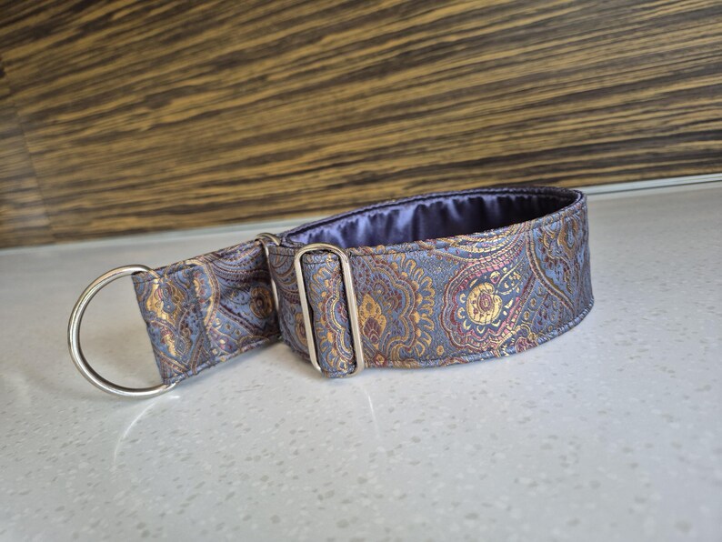 May include: A patterned dog collar with a silver metal D-ring and buckle. The collar features a paisley design in shades of blue, gold, and purple. The interior lining is a solid purple fabric. The collar is designed for a pet.