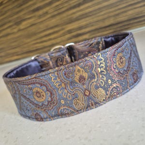May include: A patterned dog collar with a blue, gold, and burgundy paisley design. The collar has a soft, blue interior and silver metal hardware. The collar is designed for a dog.