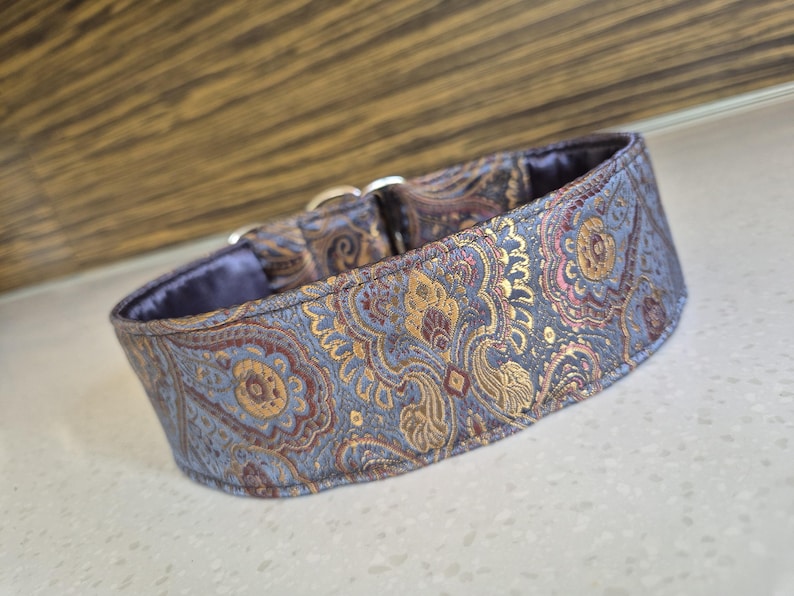 May include: A patterned dog collar with a blue, gold, and burgundy paisley design. The collar has a soft, blue interior and silver metal hardware. The collar is designed for a dog.