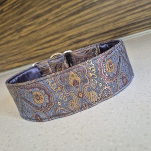 May include: A dog collar with a paisley pattern in shades of blue, gold, and burgundy. The collar has a soft, blue interior and silver metal hardware. The fabric has a subtle sheen.