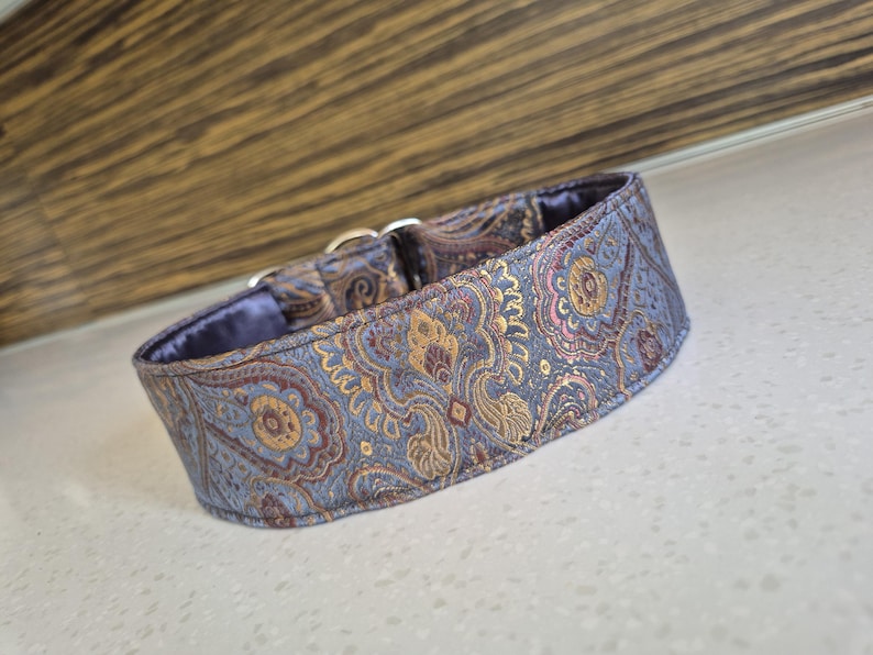 May include: A dog collar with a paisley pattern in shades of blue, gold, and burgundy. The collar has a soft, blue interior and silver metal hardware. The fabric has a subtle sheen.